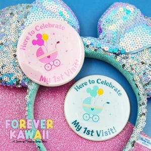 Könnte beinhalten: Zwei runde Buttons mit dem Text "Here to Celebrate My 1st Visit!" zeigen einen Kinderwagen und Mickey Mouse-förmige Ballons. Die Buttons sind rosa und hellblau, mit einem glitzernden Stirnband im Hintergrund.