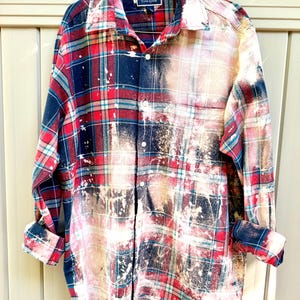 May include: A long-sleeved, button-down plaid shirt in red, navy, and white, with a bleached, distressed design. The cuffs are rolled up. The shirt is hanging on a gold hanger.
