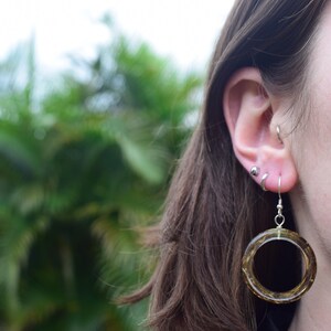 May include: A close-up of an ear with multiple piercings. A large, circular earring with a dark, textured interior hangs from the ear. The earring is suspended by a silver hook. The background is a blurred view of green foliage.