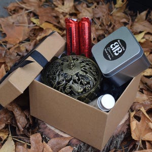 Puede incluir: Una caja de regalo con una cinta negra contiene un quemador de incienso decorativo de metal, dos tubos rojos, una pequeña lata plateada con un logotipo y un pequeño frasco. La caja está sobre hojas de otoño.