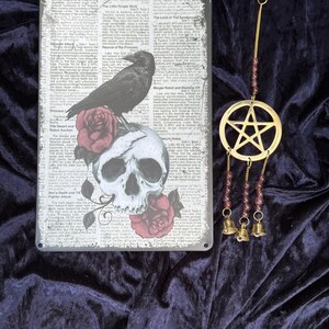 Pode incluir: Uma placa de metal com um corvo empoleirado em um crânio e rosas, contra um fundo de jornal. Um pingente de pentagrama dourado com sinos pendura perto. A placa é retangular e tem uma corda de juta para pendurar.