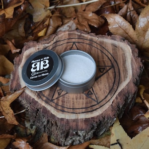 May include: A small, silver tin containing white powder. The tin has a black label with the text "Always to Help AB Dover to Harm". The tin is resting on a wooden surface with a pentagram design.
