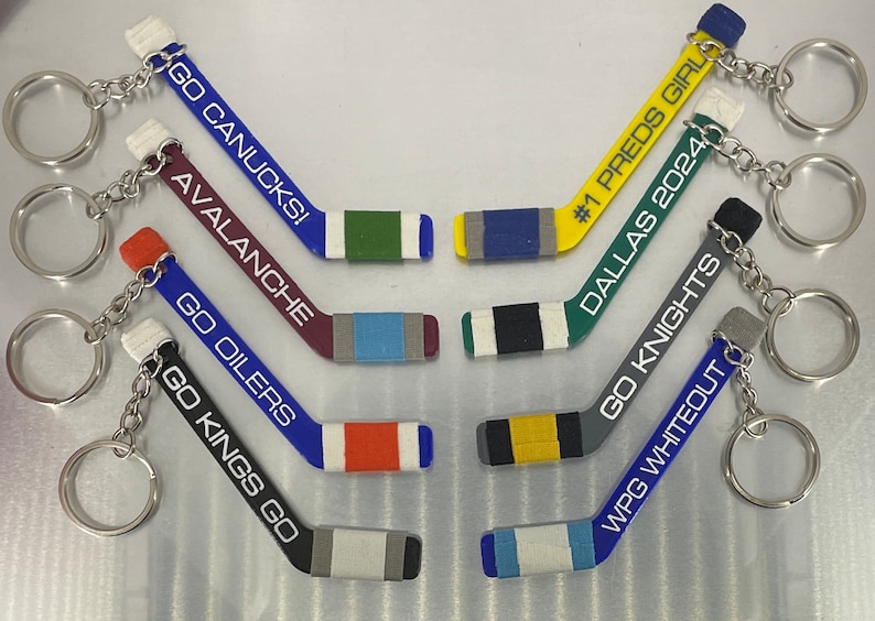 May include: A set of ten hockey stick keychains with different team logos and names. The keychains are made of plastic and have a silver metal key ring. The team names include "Go Canucks!", "Avalanche", "Go Oilers", "Go Kings Go", "#1 Preds Girl", "Dallas 2024", "Go Knights", and "WPG Whiteout".
