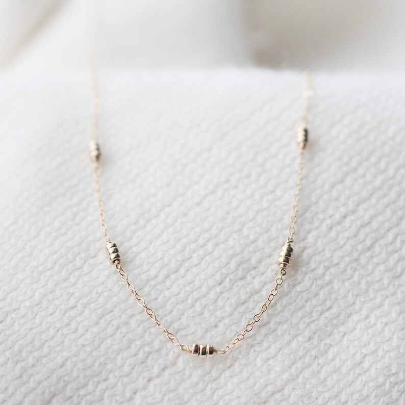 May include: A delicate gold chain necklace with small, stacked bead accents. The necklace is on a white textured surface.