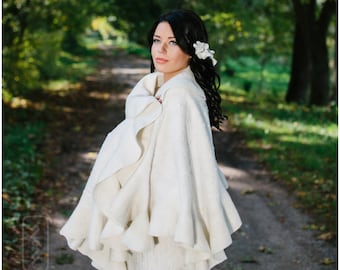 Pode incluir: Uma capa de lã branca com uma borda franjada. A capa está sendo usada por uma mulher com cabelo escuro e uma flor branca no cabelo. A mulher está em um cenário de floresta.