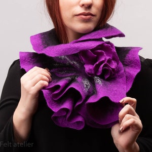 May include: A purple felted wool scarf with a ruffled design and a flower accent. The scarf is worn around the neck of a person.