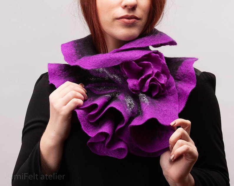 May include: A purple felted wool scarf with a ruffled design and a flower accent. The scarf is worn around the neck of a person.