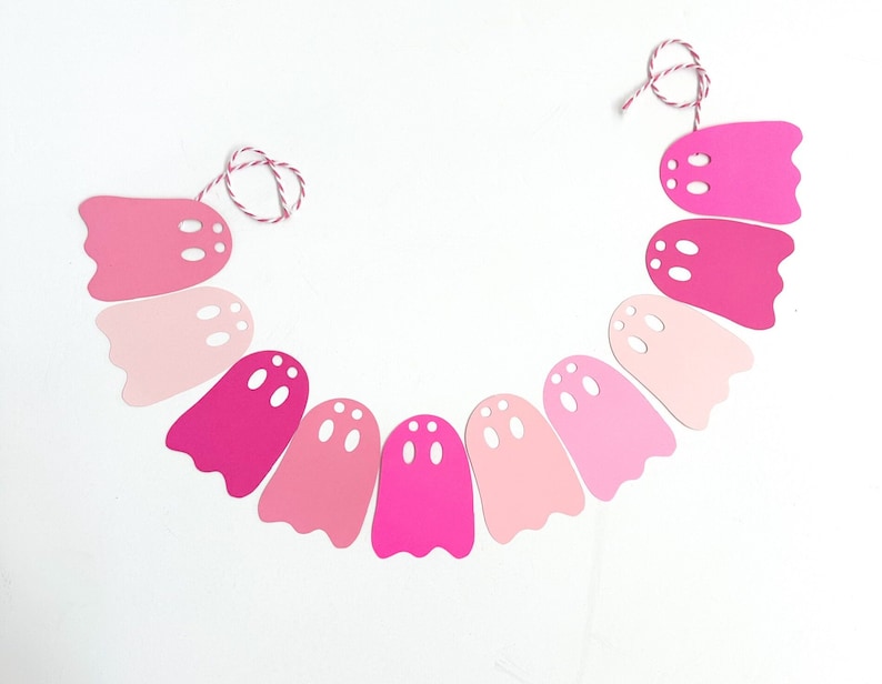 May include: A pink ghost-shaped garland with varying shades of pink. Each ghost has three eye holes and is strung together with a red and white twine. Perfect for Halloween or a themed party.