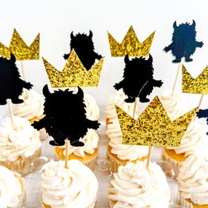 May include: Cupcakes with white frosting, topped with black monster silhouettes and gold glitter crowns on wooden sticks. The cupcakes are arranged in a clear plastic container, perfect for a birthday party or themed event.