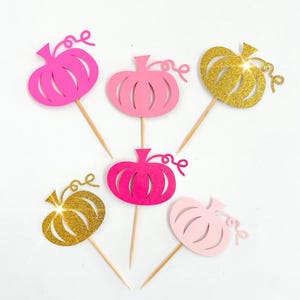 May include: Six cupcake toppers featuring pink and gold glitter pumpkins. The pumpkins have a stem and a curly vine. The toppers are perfect for a fall-themed party.
