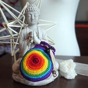 May include: A handmade crochet pouch in a rainbow color scheme, featuring concentric circles of red, orange, yellow, green, blue, and purple. The pouch has a drawstring closure and a keychain attachment. The background includes a statue of a Buddha and a white star-shaped decoration.