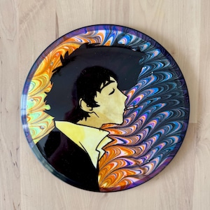 May include: Round decorative plate featuring a profile of a person with black hair and a cigarette. The plate has a black background with a colorful, swirled design in orange, blue, purple, and white. The person's silhouette is in black and gold.
