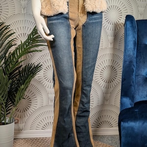 May include: Blue denim jeans with tan suede-like panels down the sides and center. The jeans have a flared leg and are displayed on a mannequin. A beige faux fur vest is worn on top.