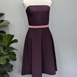 Puede incluir: Un vestido sin tirantes, de largo hasta la rodilla, de color morado oscuro con una cinta de terciopelo rosa alrededor de la cintura.