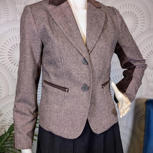 Peut inclure: Un blazer en tweed marron avec un col et des coudières en velours marron foncé. Le blazer a deux poches avant zippées et deux boutons. Il est associé à une jupe plissée noire.
