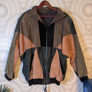 Peut inclure: Une veste en daim à blocs de couleurs présentant un motif géométrique dans des tons vert olive, beige et noir. La veste est dotée d'une fermeture éclair frontale intégrale, d'un col montant, de manches longues avec des poignets côtelés noirs.