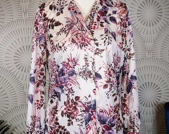 Puede incluir: Camisa abotonada blanca con estampado floral en tonos morados y burdeos. La camisa de manga larga se exhibe en un maniquí. El diseño floral incluye hojas y flores.