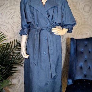May include: A long, double-breasted trench coat in a muted blue shade with a textured finish. The coat features a wide notched collar, long sleeves with cuffs, a matching tie belt at the waist, and a series of round buttons down the front.