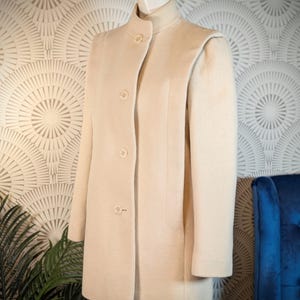 May include: A cream-colored coat with a high collar and button closure. The coat is displayed on a mannequin, showcasing its tailored design and shoulder detailing. The coat is knee-length and appears to be made of a soft, wool-like material.