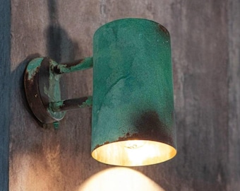 May include: A green metal wall sconce with a cylindrical shade and a light bulb that is turned on. The light casts a warm glow on the wall.