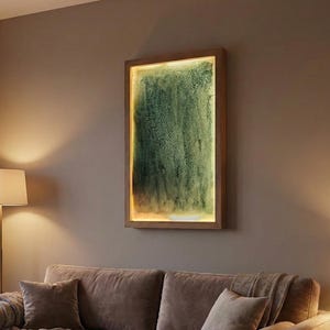 Puede incluir: Un salón con un sofá marrón, cojines y mantas. Una mesa de centro de madera con una taza y libros. Sobre el sofá, una obra de arte enmarcada con un centro texturizado verde y un borde de luz cálida.