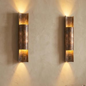 May include: Two vertical, copper-colored wall sconces with a textured finish are mounted on a beige wall. Each sconce has three sections, with the middle section being a long rectangle. Warm light emanates from the top and bottom sections.
