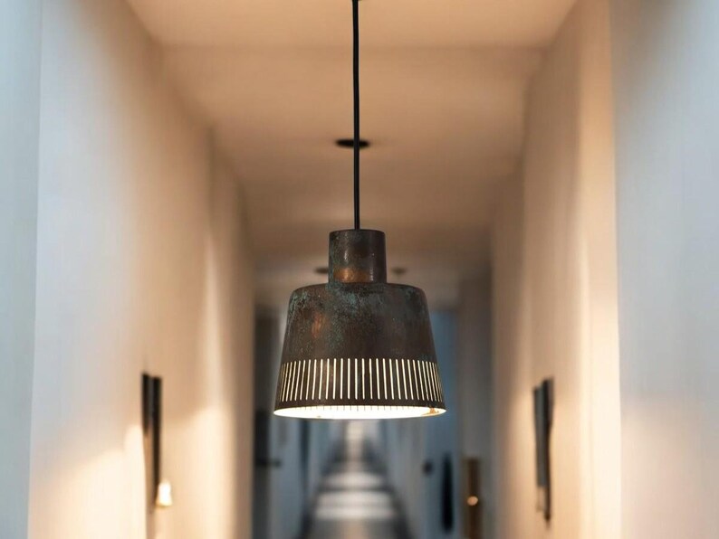 May include: A black metal pendant light fixture with a copper-colored shade featuring a geometric pattern of cut-outs. The light is hanging from a white ceiling in a hallway.