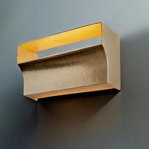 May include: A wall-mounted light fixture with a curved, gold-toned metal frame and a rectangular, frosted glass panel. The light is turned on, illuminating the glass panel with a warm, yellow glow.