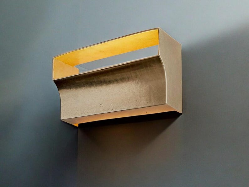 May include: A wall-mounted light fixture with a curved, gold-toned metal frame and a rectangular, frosted glass panel. The light is turned on, illuminating the glass panel with a warm, yellow glow.