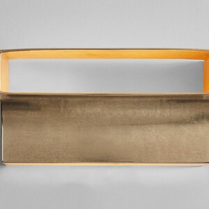 May include: A gold-toned rectangular wall sconce with a slightly curved bottom edge. The sconce has a thin, rectangular opening at the top.