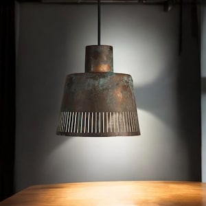 May include: A copper pendant light with a perforated shade hangs from the ceiling above a wooden table. The light is on and casting a warm glow.