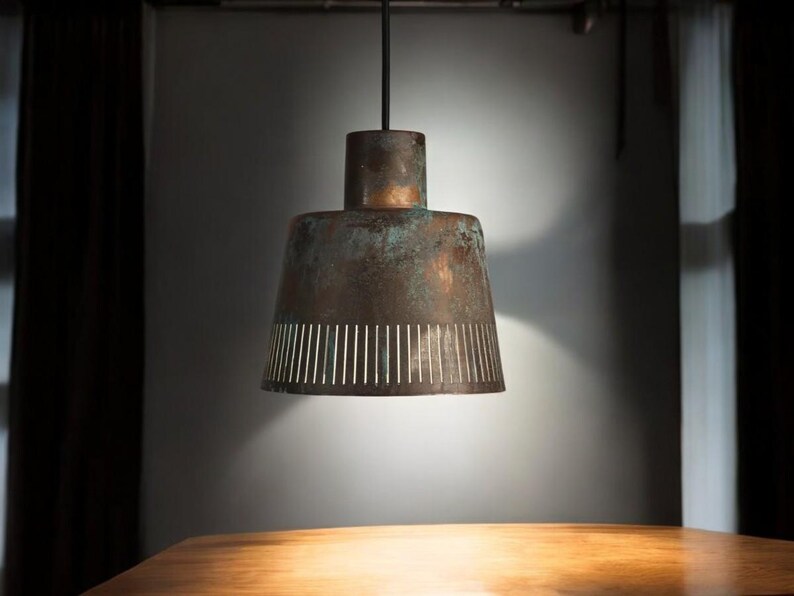 May include: A copper pendant light with a perforated shade hangs from the ceiling above a wooden table. The light is on and casting a warm glow.