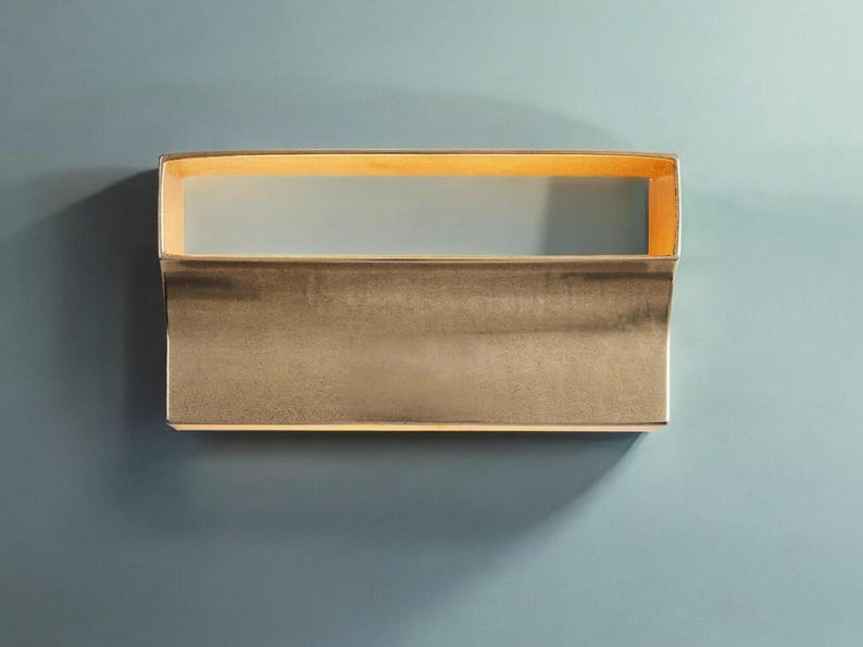 May include: A gold-toned wall sconce with a rectangular shape and a curved bottom edge. The sconce has a light inside that illuminates the room.