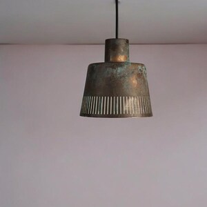 May include: A copper-toned pendant light with a perforated design hangs from a black cord against a light pink wall.