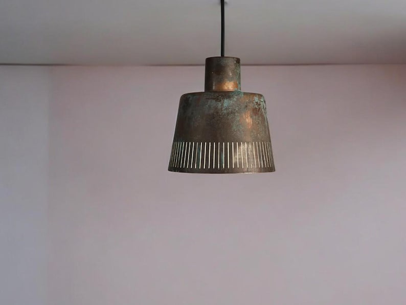 May include: A copper-toned pendant light with a perforated design hangs from a black cord against a light pink wall.