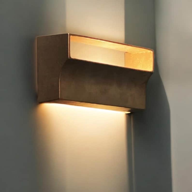 May include: A modern wall sconce with a rectangular shape and a warm, golden finish. The light fixture has a built-in LED light that casts a soft glow.