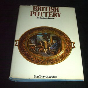 May include: A book titled "British Pottery: An Illustrated Guide" by Geoffrey A. Godden. The cover features a colorful illustration of a ceramic plate with a scene of people and dogs in a rustic setting.