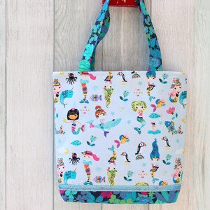 May include: A light blue tote bag with a colorful mermaid and sea creature pattern. The bag has blue floral handles and a blue trim at the bottom. The bag is hanging against a light wood background.