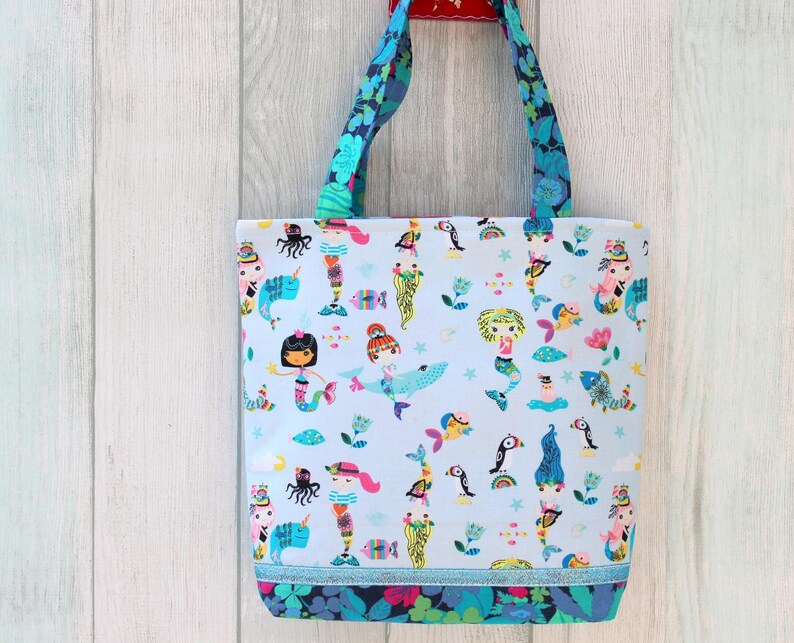 May include: A light blue tote bag with a colorful mermaid and sea creature pattern. The bag has blue floral handles and a blue trim at the bottom. The bag is hanging against a light wood background.