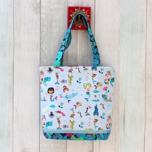May include: A light blue tote bag with a colorful mermaid and sea creature pattern. The bag has blue floral handles and a matching floral trim at the bottom. The bag is hanging on a white wooden wall.