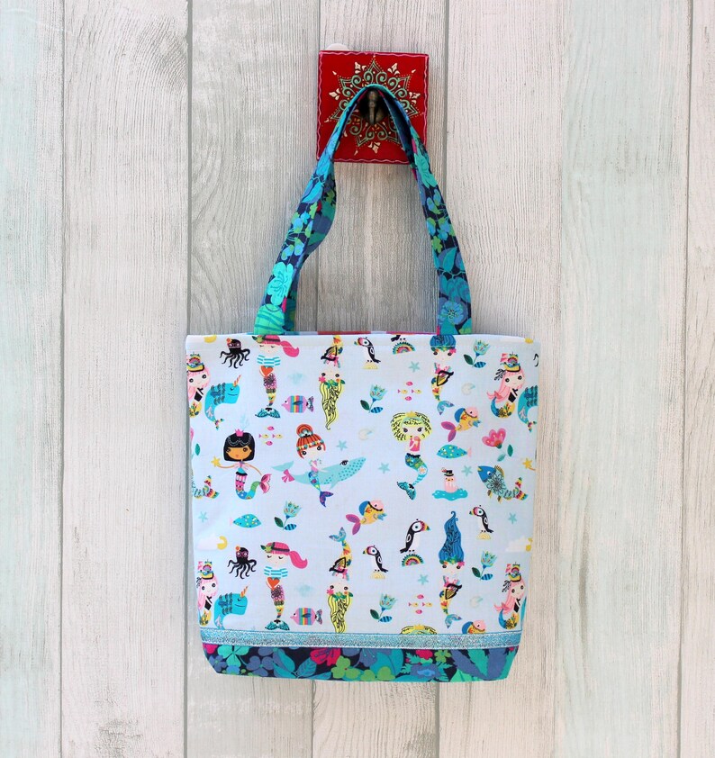 May include: A light blue tote bag with a colorful mermaid and sea creature pattern. The bag has blue floral handles and a matching floral trim at the bottom. The bag is hanging on a white wooden wall.