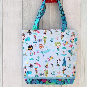 May include: A light blue tote bag with a colorful mermaid and sea creature pattern. The bag has blue floral handles and a matching trim at the bottom. The bag is hanging against a light wood background.