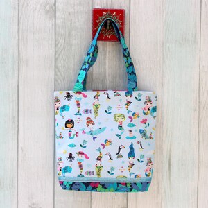May include: A light blue tote bag with a colorful mermaid pattern. The bag features blue floral handles and a matching floral trim at the bottom. The design includes various mermaids, sea creatures, and aquatic plants. The bag is hanging against a white wooden background.
