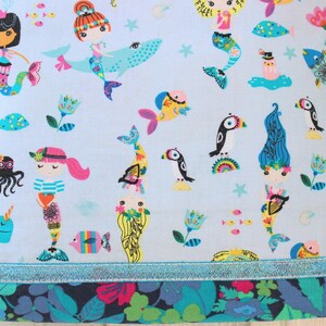 May include: Light blue fabric with a whimsical mermaid and sea creature pattern. The design includes mermaids, fish, octopuses, and other marine life in bright colors. A floral border and a blue ribbon trim the bottom.
