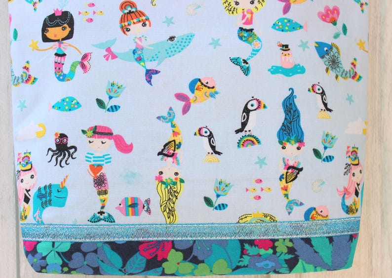 May include: Light blue fabric with a whimsical mermaid and sea creature pattern. The design includes mermaids, fish, octopuses, and other marine life in bright colors. A floral border and a blue ribbon trim the bottom.