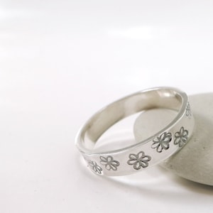 May include: A silver band ring with a row of small flower designs around the band.
