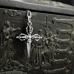 May include: A silver pendant in the shape of a cross with a dagger-like point. The cross has intricate details and a crown at the top.
