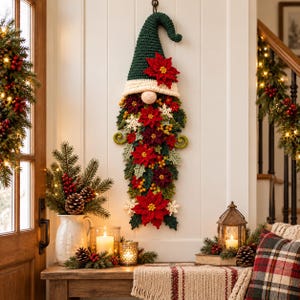 May include: A wall-hanging crochet gnome decoration featuring a tall dark green pointed hat, a cream-coloured brim, and a round nose. The beard is composed of layered red and dark red poinsettia flowers, green leaves, small yellow berries, and white snowflakes.
