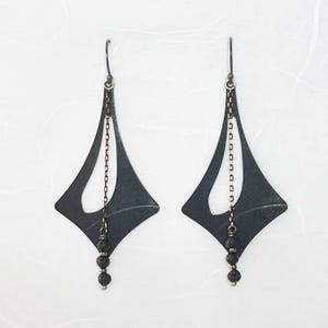 May include: A pair of black geometric earrings with a diamond shape. The earrings have a chain and three black beads hanging from the bottom. The ear wires and chain accents are gold-colored.