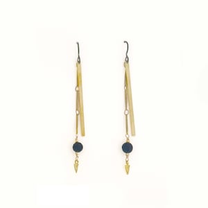 May include: A pair of dangling earrings with a minimalist design. Each earring features a long, rectangular gold-colored bar, connected by chains to a small, dark blue bead and a gold-colored arrowhead pendant. The earrings hang from a dark metal hook.